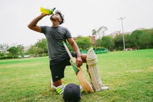 Full length of ethnic male cricket player in sportswear with bat and helmet and pads kneeling on grass while drinking water from sport bottle